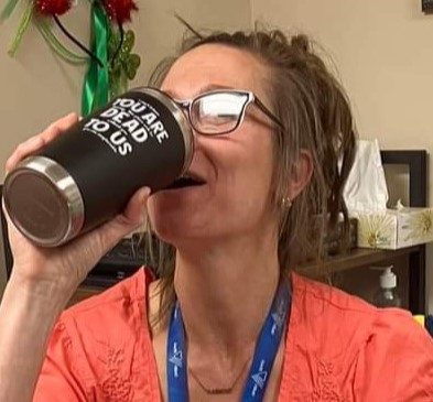 A woman with light brown hair pulled back in a bun drinks from a traveller mug. She's wearing a pink shirt, a blue lanyard, and glasses.