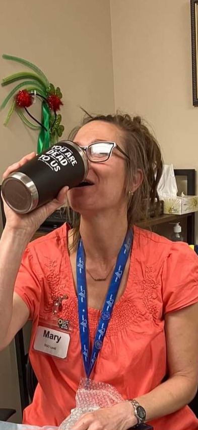 A woman with light brown hair pulled back in a bun drinks from a traveller mug. She's wearing a pink shirt, a blue lanyard, and glasses.