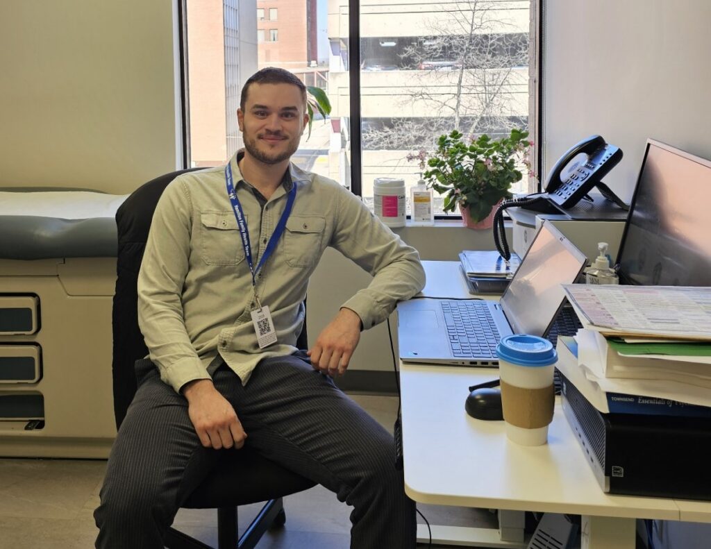 A man with short cropped hair and a short beard sits in an office chair in a clinical room and smiles at the camera. He is wearing black pants, a light grey collared shirt, and a blue lanyard.