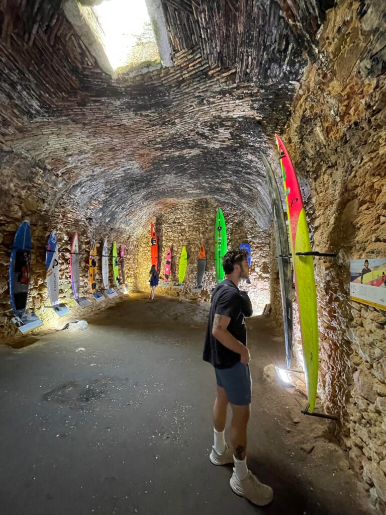 A man stands in a cave and looks at surfboards that are mounted on the wall of the cave.