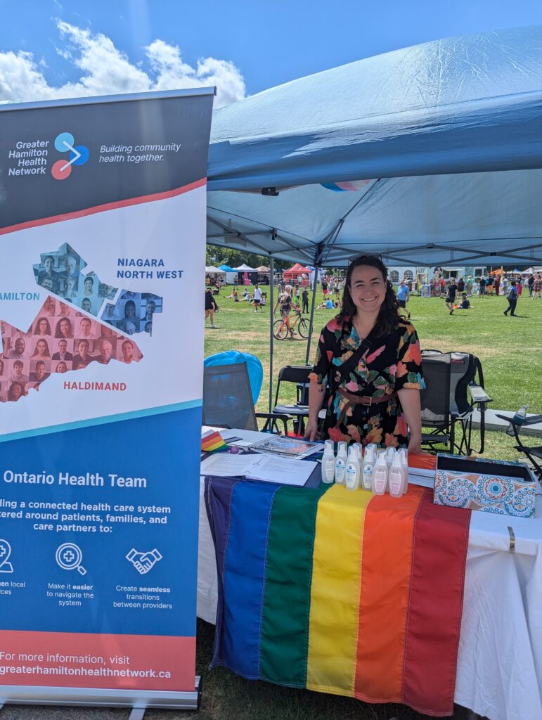 A woman with long curly brown hair smiles at the camera. She is wearing a jumpsuit with rainbow flowers on it. She is standing under a tent behind a table with a Pride flag and a sign next to her that says "Greater Hamilton Health Network".