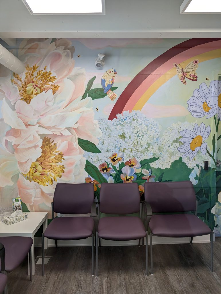 The Eva Rothwell waiting room with a wall mural of flowers, rainbows and insects with chairs set up and a side table.