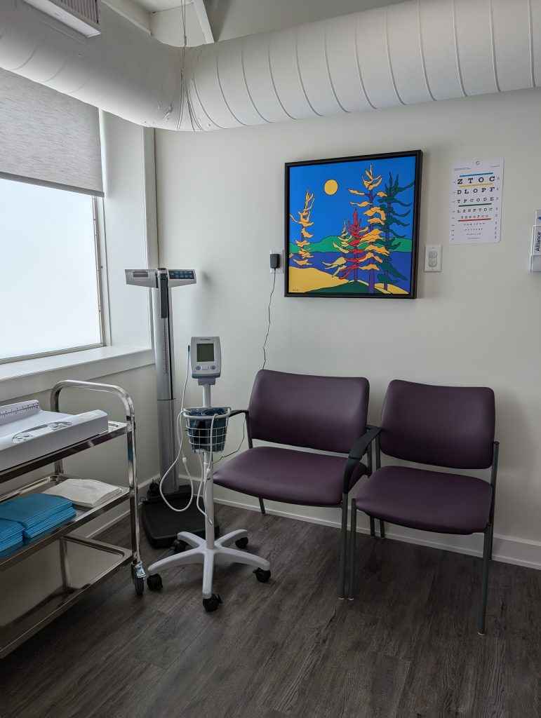 A room with shelving, scale, maching and two chairs with a painting of trees and a sky on the wall.