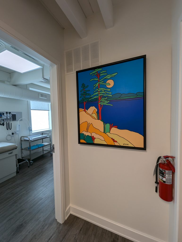 A hallway with a painting of trees hanging on the wall, with a door open, show the inside of a room with a window.