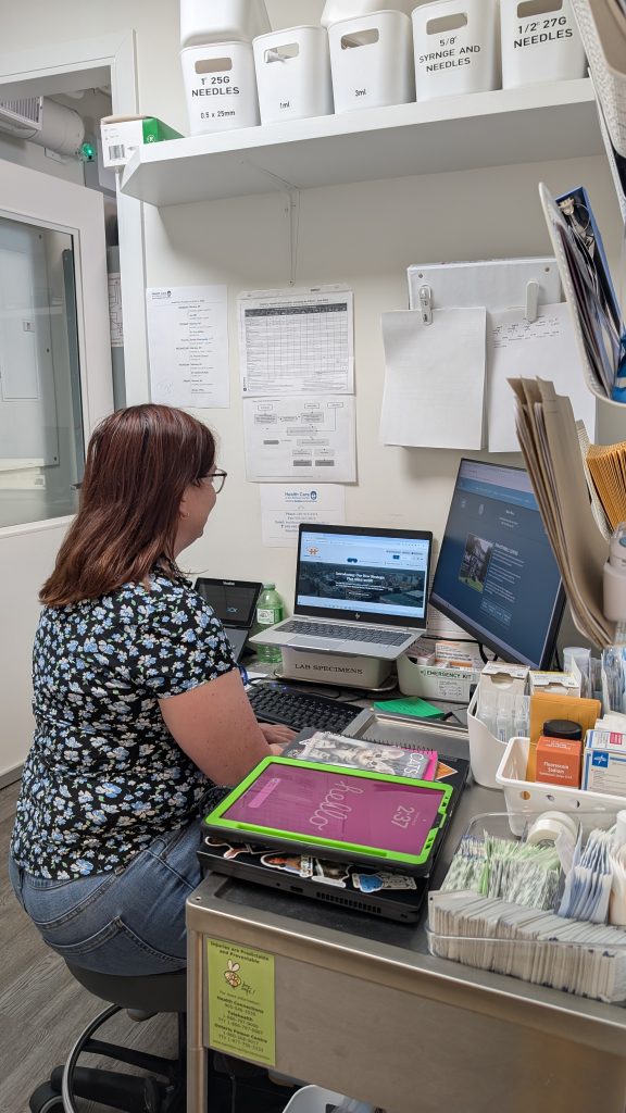 Someone at their desk typing on a computer looking at two screens, with healthcare related items around the desk and on shevles.