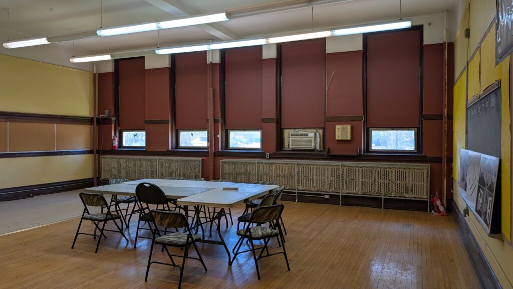 An old classroom with yellow walls, small windows, wood floors and a table in the middle.