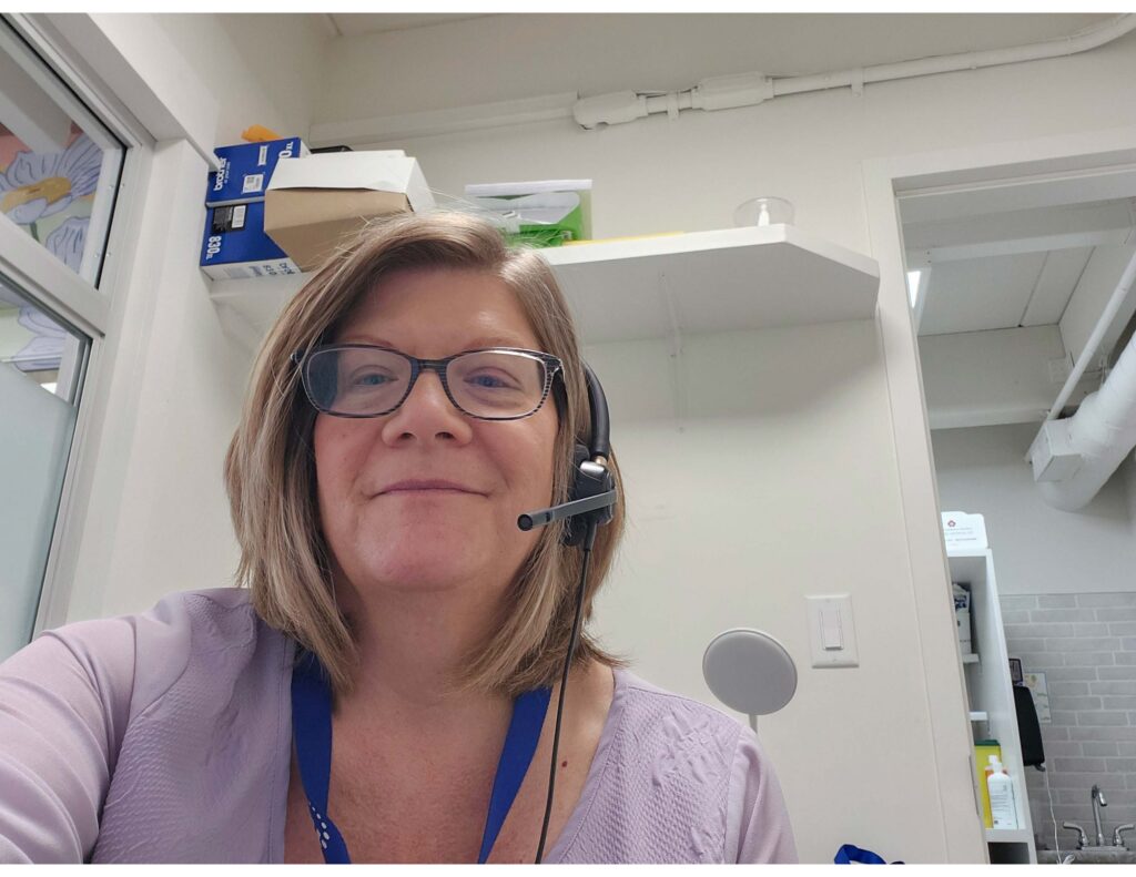 A woman with short light hair and glasses smiles at the camera while she sits at a clinic's front desk. She's wearing a headset.