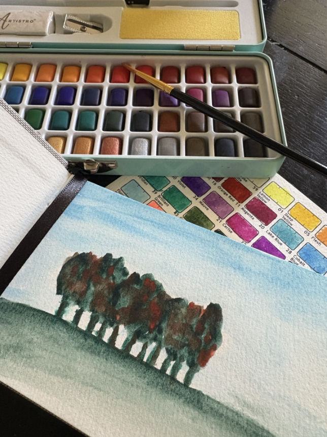A watercolour pallete and paintbrush sits next to a sketchbook with a lanscape painting of trees.