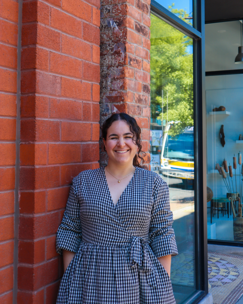 A woman with brown curly hair stands in front of a red brick wall and the glass windows of a shop and smiles at the camera. She is wearing a black and white checkered dress.