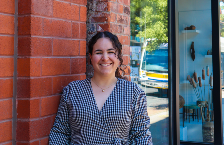 A woman with brown curly hair stands in front of a red brick wall and the glass windows of a shop and smiles at the camera. She is wearing a black and white checkered dress.