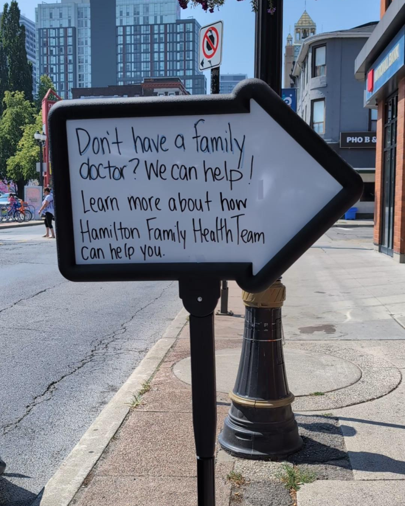 An arrow-shaped whiteboard sign that says "Don't have a family doctor? We can help! Learn more about how the Hamilton Family Health Team can help you."