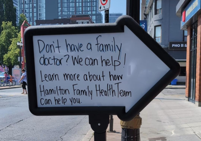 An arrow-shaped whiteboard sign that says "Don't have a family doctor? We can help! Learn more about how the Hamilton Family Health Team can help you."