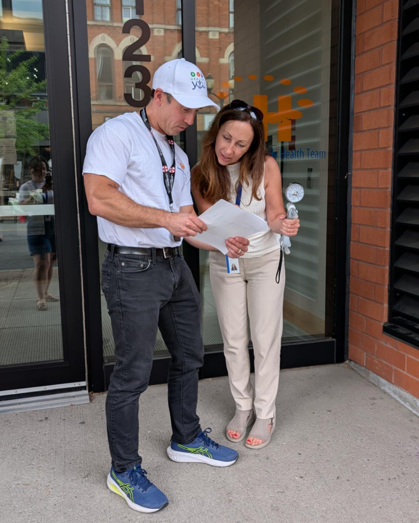 A man and a woman stand in front of an office building and look at a piece of paper. The woman is holding a machine that tests grip strength.