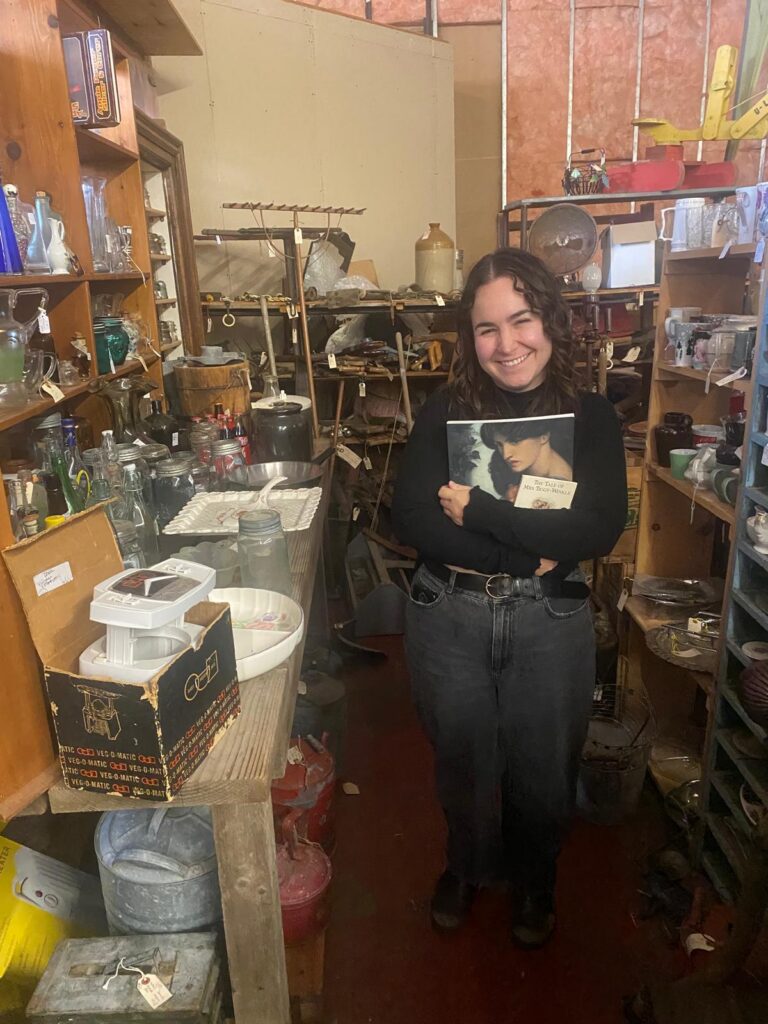 A woman with long brown curly hair stands in an antique mall and smiles at the camera. She is holding two books. She is wearing a black long-sleeved shirt and black jeans.